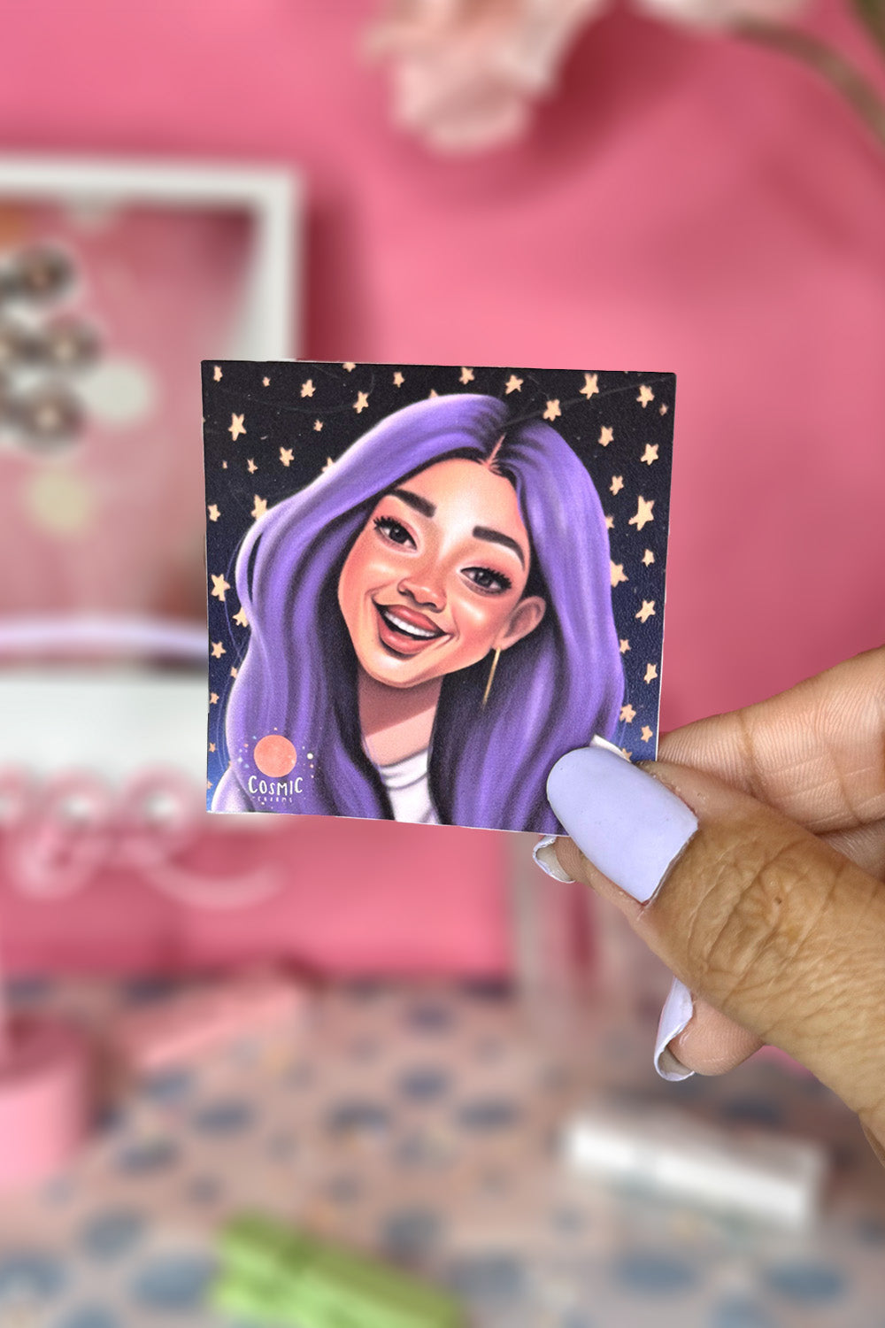 Hand holding a square sticker of Celeste Estrella smiling with long pastel purple hair on a black starry background.